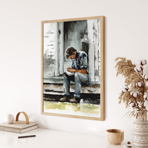 Watercolor painting of a young man praying on a porch in a light wood frame hanging slightly above a white colored surface that has a neutral colored book, cup, and a neutral vase with cotton flower buds and dried grass