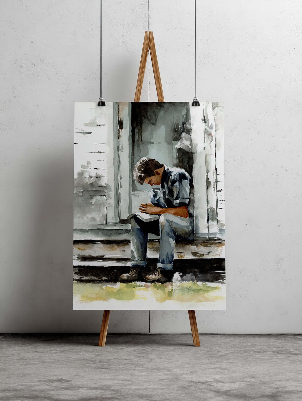 watercolor painting of a young man praying on a porch of a white house with gray shadows and light hitting the house and door behind him painting is large and sitting on a wood colored easel against a light colored wall and sitting on light gray carpet 